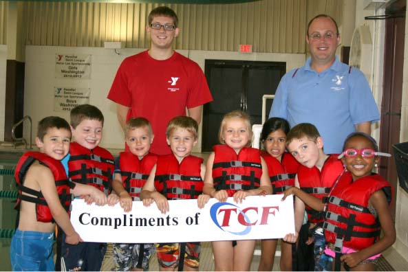 Group of young children who received a grant from the community's foundation hold a sign that reads, "compliments of TCF," while smiling.