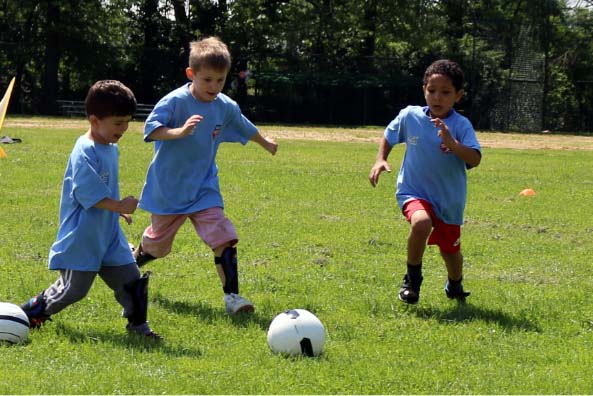 Three young children play run toward a soccer ball on a sunny day.