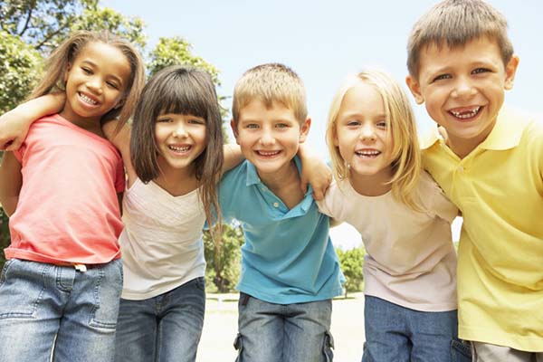Five children lean toward the camera smiling with their arms around each other's shoulders.